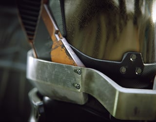 Close-up view of a worn, metallic and carbon-fiber armor piece with bolts and orange accents, suggesting protective gear or a futuristic helmet.